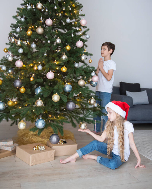 Παιδιά - ένα αγόρι και ένα κορίτσι παίζουν κοντά στο χριστουγεννιάτικο δέντρο. Σαλόνι εσωτερικό με χριστουγεννιάτικο δέντρο και διακοσμήσεις. Πρωτοχρονιά. Δώρο. - Φωτογραφία, εικόνα