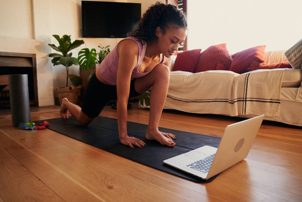 Heureuse jeune femme adulte multiculturelle suivant un tutoriel d'exercice sur son ordinateur portable à la maison dans son salon moderne - Photo, image