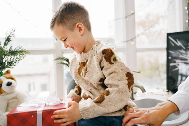 Happy little boy holding his Christmas gift sitting on windowsill - Zdjęcie, obraz