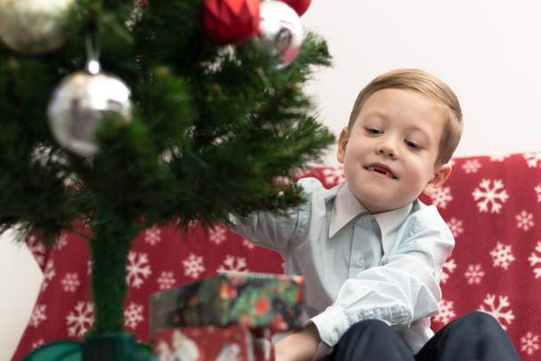 7歳の少年は、暖かい毛布の上に座っている新年の木の背景に新年の贈り物に満足しています。選択的フォーカス。肖像画 - 写真・画像