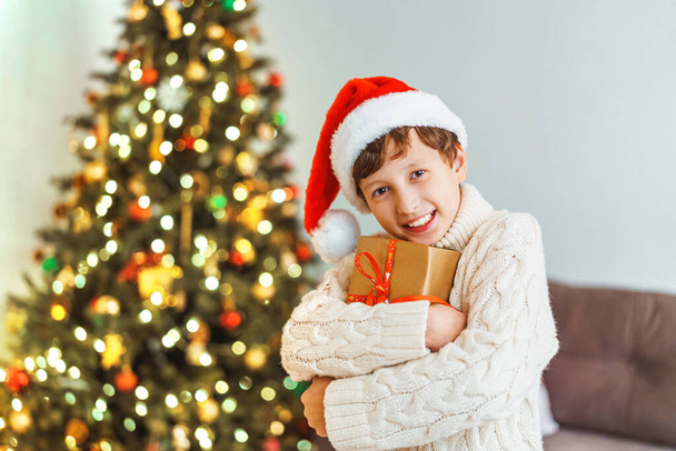 Merry Christmas. cheerful boy in knitted sweater and Santa hat, holding gift, standing against background room decorated with twinkling lights for Christmas. child laughs and enjoys the gift. - Fotó, kép