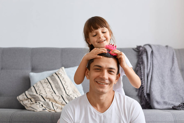 Portrait of smiling happy attractive man and his little daughter, family time and having fun together, sitting near sofa, daughter making funny hairstyle for dad, expressing positive emotions. - Photo, image