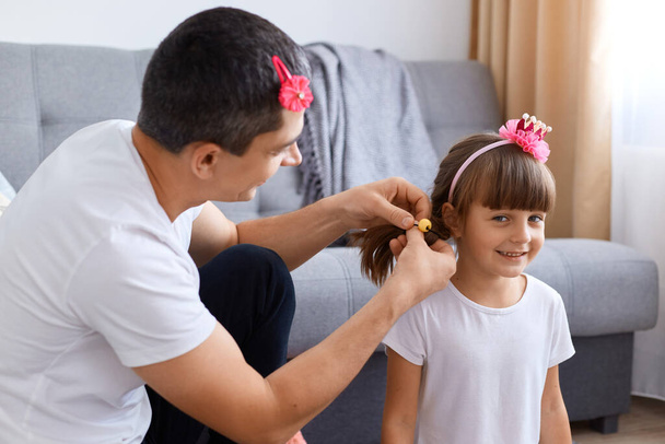 Portrait of brunette man spending time with his daughter, making pigtails while sitting on floor near sofa, smiling cute girl with charming smile looking at camera. - Valokuva, kuva