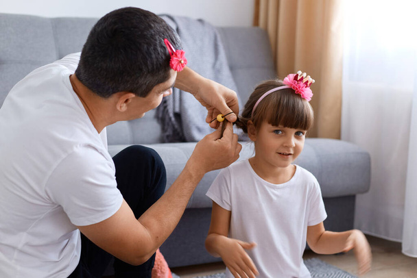 Indoor shot of happy family sitting on floor at home in living room, father braiding girl's hair, family time at weekend, happy child with hie lovely daddy. - Fotoğraf, Görsel