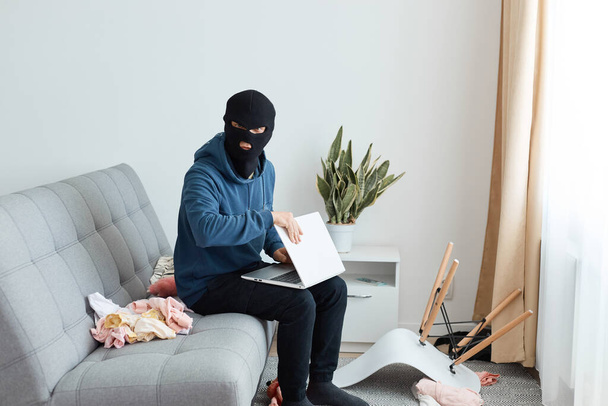 Indoor shot of male burglar wearing dark blue hoodie and black mask entered someone else's house and tries to steal a laptop, sits on the couch and looks at the camera. - Фото, изображение