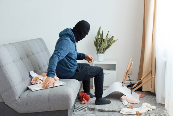 The thief found a laptop in the house which trying to rob, sits on the couch and checking the nightstand, looking for something else to steal, man robber wearing blue hoodie and black balaclava - Foto, Bild