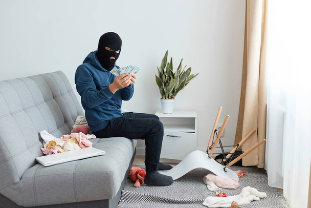 Portrait of amazed surprised man robber wearing blue hoodie and black balaclava sitting on sofa in house, being shocked to find big sum of money, expressing astonishment. - Фото, изображение
