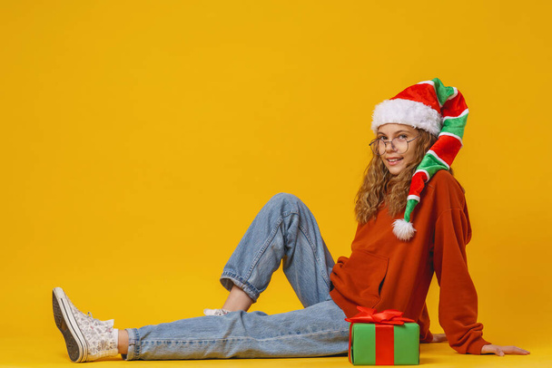 little surprised girl in Santa gnome hat holds a Christmas gift box sit on yellow background in studio. child laughs and receives a lot of gifts for Christmas advertising. - Фото, зображення