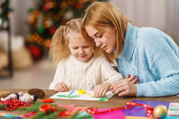 cute little girl and her mother are engaged in needlework, creativity, enjoying the process, sitting at a desk, a lot of materials, preparing for a holiday in a decorated house. - Foto, immagini