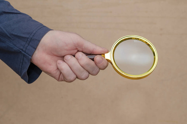 Ein erwachsener Mann hält vor braunem Hintergrund eine Lupe in der Hand. Eine große runde Lupe in einem goldenen Rahmen. Nahaufnahme. Selektiver Fokus. - Foto, Bild