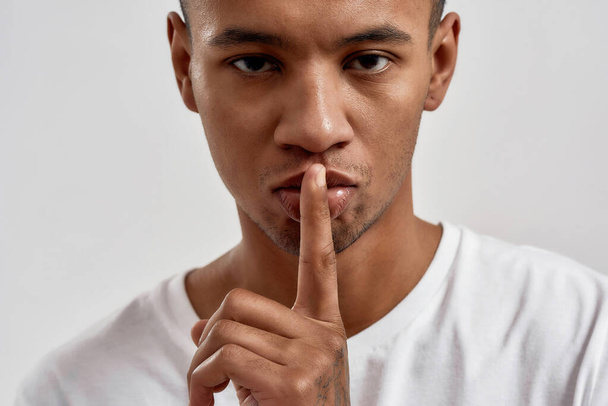 Portrait of dark skinned young guy showing shh gesture, keeping a secret, posing isolated over white background - Fotografie, Obrázek