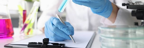 Mujer científica sostiene tubo de ensayo con líquido azul en guantes y escribe los datos químicos obtenidos - Foto, imagen