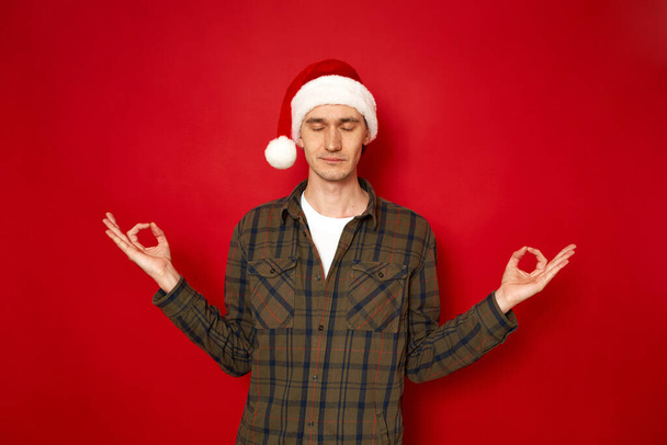 rauhallinen valkoihoinen mies meditoi sisätiloissa, pitää kädet mudan eleessä, silmät kiinni, yrittää rentoutua pitkien jouluvalmistelujen jälkeen, pitää sormia joogan merkissä, eristettynä punaiseen seinään - Valokuva, kuva
