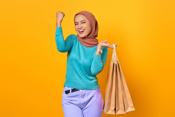 Возбужденная и веселая молодая азиатка держит сумки с покупками на желтом фоне - Фото, изображение