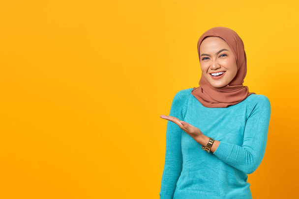 Portrait of smiling young Asian woman showing copy space on palm isolated over yellow background - Φωτογραφία, εικόνα