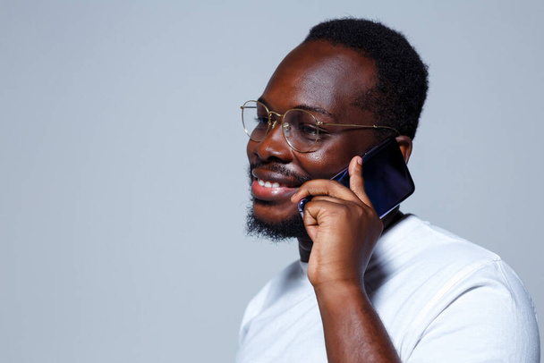 Un uomo africano che tiene un telefono blu nella mano sinistra e sorride. Indossa una maglietta bianca e occhiali.. - Foto, immagini
