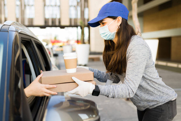 Delivering food during the pandemic. Young woman with a face mask giving the pizza order to a customer in the car  - Photo, Image