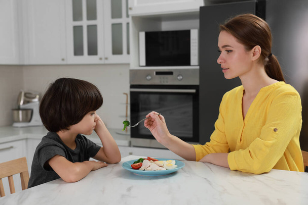 Mother feeding her son in kitchen. Little boy refusing to eat dinner - Zdjęcie, obraz
