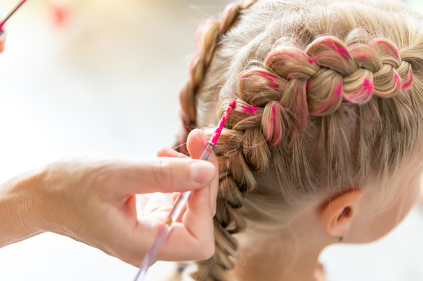 小さな娘の子供の頭の上にピンクの色で染めの組紐を作るブラシを保持母の手を閉じます。若い女の子にキラキラペイント編組髪を適用美容師スタイリスト。パーティーのお祝いを - 写真・画像