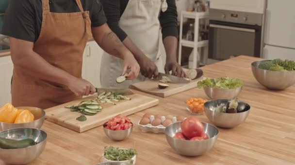 Tilting-up of young African American female and male sous-chefs wearing aprons, standing at kitchen island, cutting fresh cucumber and eggplant on cutting boards - Footage, Video