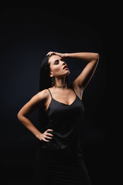 brunette young woman in slip dress posing with hand on hip isolated on black  - Fotó, kép