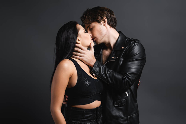 man in leather jacket kissing sexy woman in black crop top on grey - Фото, зображення