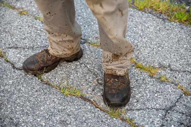 Un homme en bottes et pantalons de travail éclaboussés sur une rue asphaltée cassée - Photo, image