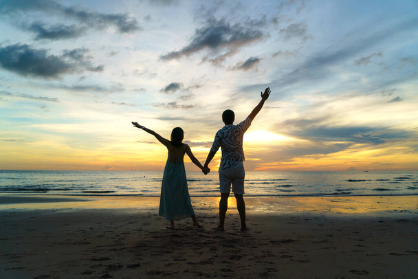 Romantik Asyalı çift ayakta ve el ele tutuşup tatil boyunca sahilde gün batımının tadını çıkarıyorlar.. - Fotoğraf, Görsel
