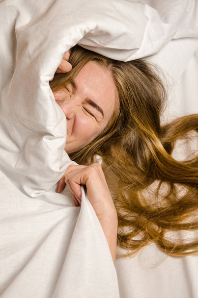 Όμορφη νεαρή γυναίκα ξυπνά το πρωί στο λευκό κρεβάτι στις ακτίνες του ήλιου, έχει πλούσια κυματιστά ξανθά, χρυσά μαλλιά - Φωτογραφία, εικόνα