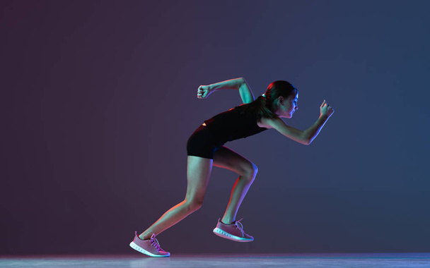 Full-length side view portret dziewczyny, profesjonalny sportowiec, trening biegowy odizolowany ponad gradientowym niebieskim fioletowym tle w neonie - Zdjęcie, obraz