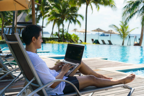 Asiatische Mann verbrachte seinen Sommerurlaub mit der Arbeit an seinem Laptop in einem Stuhl in der Nähe des Swimmingpools in Resort-Hotel am Meer - Foto, Bild