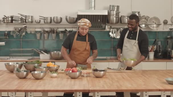 Mittlere Länge junger afroamerikanischer Sous-Köche mit Schürzen, die auf der Kücheninsel stehen, frische Gurken und Auberginen auf Schneidebrettern schneiden, reden und lächeln - Filmmaterial, Video