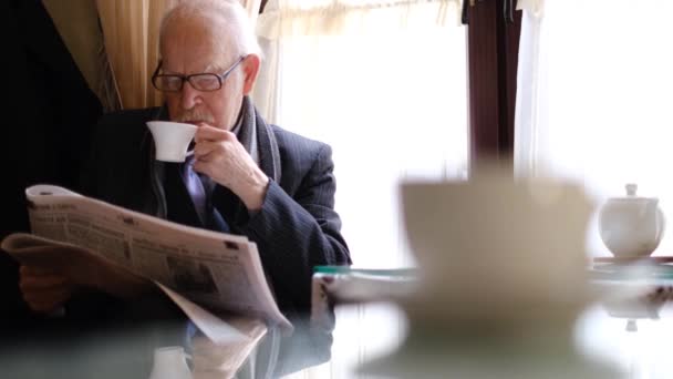 Ένας ηλικιωμένος κάθεται σε μια καρέκλα και διαβάζει εφημερίδα πάνω από τον πρωινό καφέ σε ένα καφέ.. - Πλάνα, βίντεο