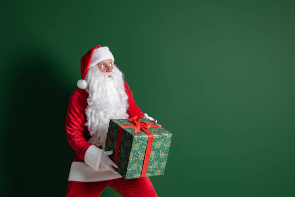 Emotional Santa Claus sneaking while holding big green gift box in hands on green studio background - Foto, afbeelding