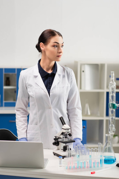 Scientifique en blouse blanche debout à proximité de matériel médical, ordinateur portable et vaccin en laboratoire  - Photo, image
