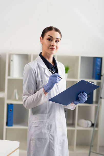 Scientist in white coat looking at camera while holding pen and clipboard in lab  - Foto, Imagen