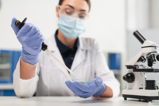 Petriho miska a elektronická pipeta v rukou rozmazaného vědce v lékařské masce v laboratoři - Fotografie, Obrázek