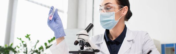 Cientista em luva de látex e máscara médica segurando tubo de ensaio perto do microscópio em laboratório, banner  - Foto, Imagem