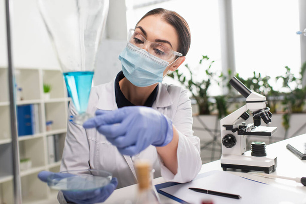 Cientista em óculos de trabalho com placa de Petri e frasco perto do microscópio em laboratório  - Foto, Imagem