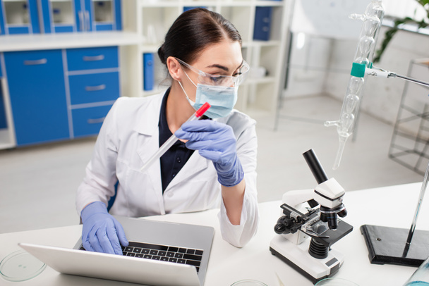 Scientist in latex gloves holding pcr test and using laptop near microscope in lab  - Photo, Image