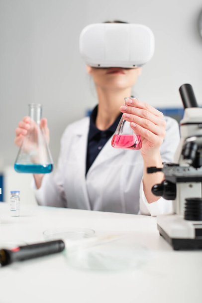 Frasco en mano del científico borroso cerca del microscopio, la vacuna y la pipeta electrónica en el laboratorio  - Foto, Imagen