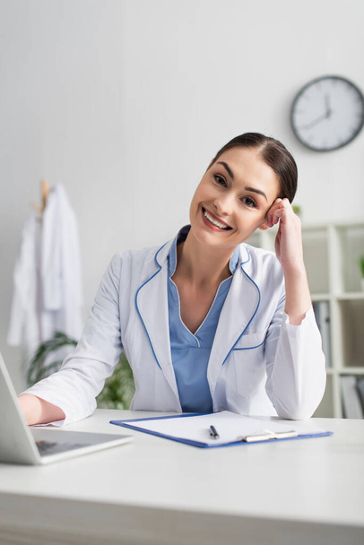 Positive doctor looking at camera near laptop and clipboard in clinic  - Foto, Imagem