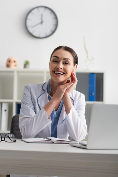 happy doctor looking at camera while sitting near blurred laptop - Photo, Image