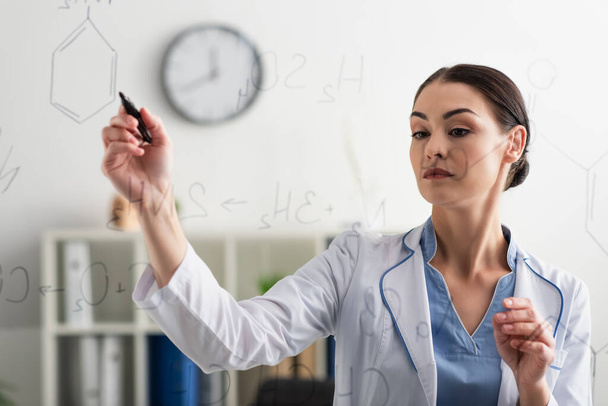 brunette doctor in white coat writing chemical formulas on glass board in lab - Foto, afbeelding