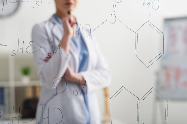 partial view of blurred doctor standing near glass board with chemical formulas - Foto, Bild