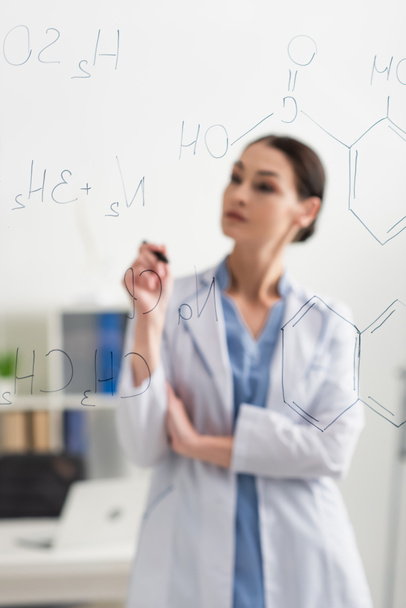 blurred doctor in white coat standing near chemical formulas on glass board - Photo, Image