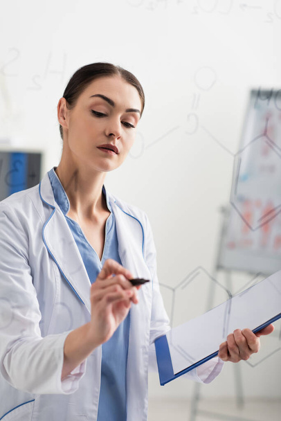 brunette chemist holding clipboard while writing formulas on glass board - Photo, Image