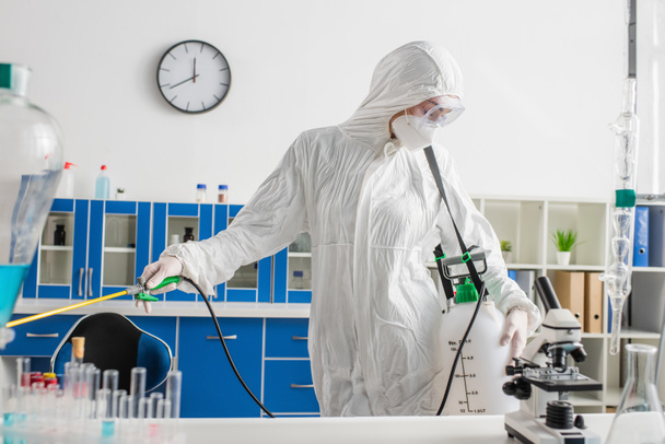 Wissenschaftler im Schutzanzug bei der Desinfektion von Laboratorien in der Nähe von Mikroskop und Reagenzgläsern auf dem Schreibtisch - Foto, Bild