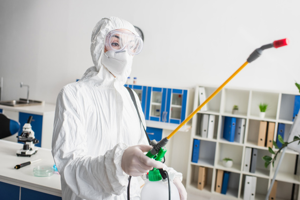 lékař v brýlích a ochranném obleku drží postřikovač při dezinfekci v laboratoři - Fotografie, Obrázek