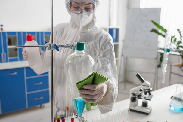 Wissenschaftler im Schutzanzug wischt Fläschchen mit Lappen ab, während er Labordesinfektion macht - Foto, Bild